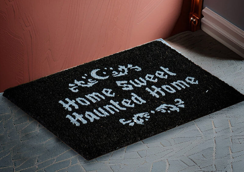 A black rectangular doormat with white gothic-style text reading 'This City Home Sweet Home Haunted Home' placed on a gray patterned floor against a pink wall.
