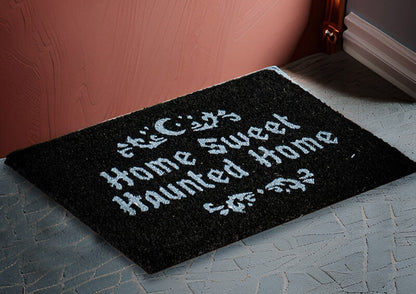 A black rectangular doormat with white gothic-style text reading 'This City Home Sweet Home Haunted Home' placed on a gray patterned floor against a pink wall.