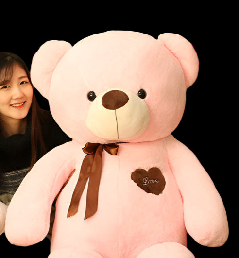 Large pink teddy bear with a heart and bow tie next to a person sitting on the floor.