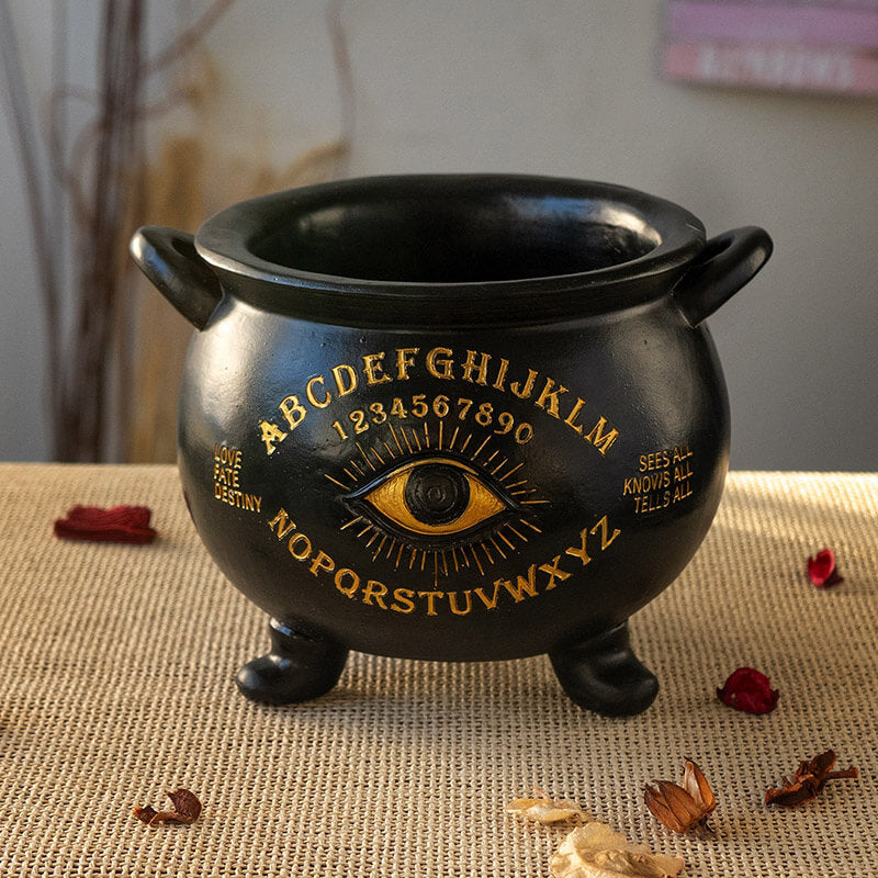 Black cauldron with gold lettering and eye design on a textured surface.