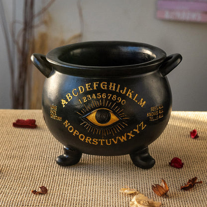 Black cauldron with gold lettering and eye design on a textured surface.