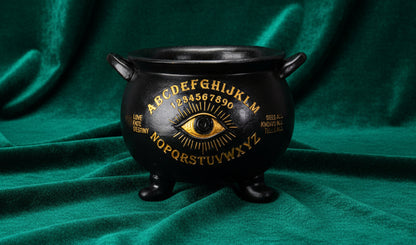 A black cauldron-shaped planchette with gold foil-style printing of an alphabet, numbers, and an eye motif, shown on a plain white background.