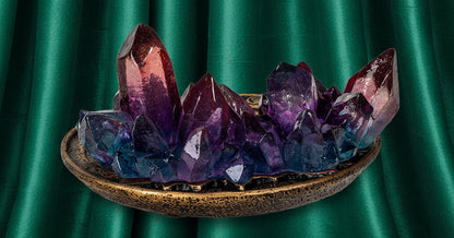 A decorative crystal cluster centerpiece featuring purple, blue, and reddish-brown amethyst-like points resting on a textured bronze-colored oval base, isolated on a white background.