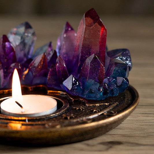 A decorative cluster of translucent crystal points in purple, blue, and red tones arranged in a bronze tray with a lit white tea light candle in front, set on a wooden surface.