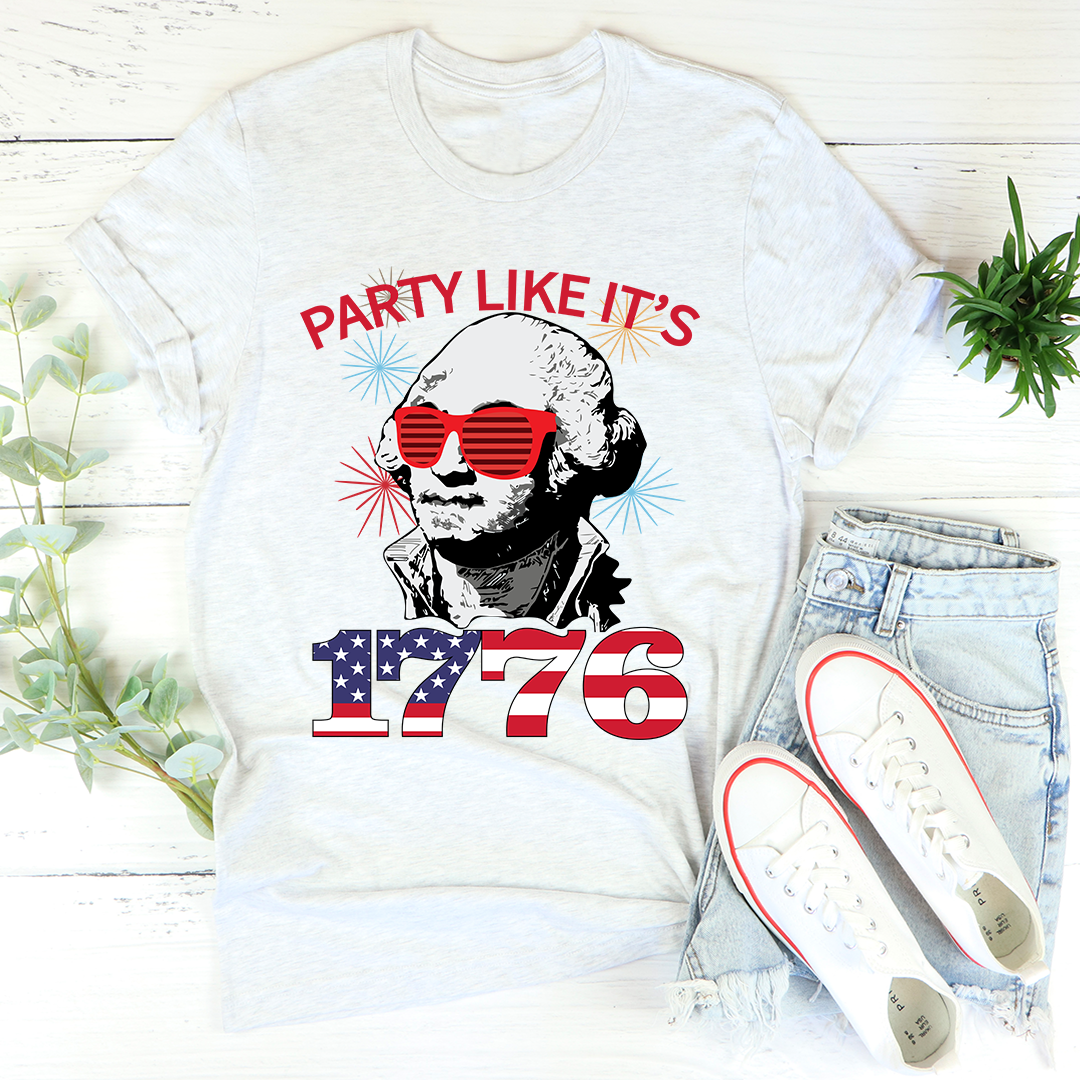 White t-shirt with 'Party Like It's 1776' design featuring a person wearing sunglasses, paired with light blue jeans and white sneakers on a wooden surface.
