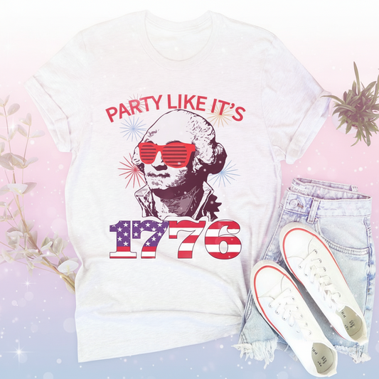 White t-shirt with 'Party Like It's 1776' design featuring a person wearing sunglasses, paired with light blue jeans and white sneakers on a wooden surface.