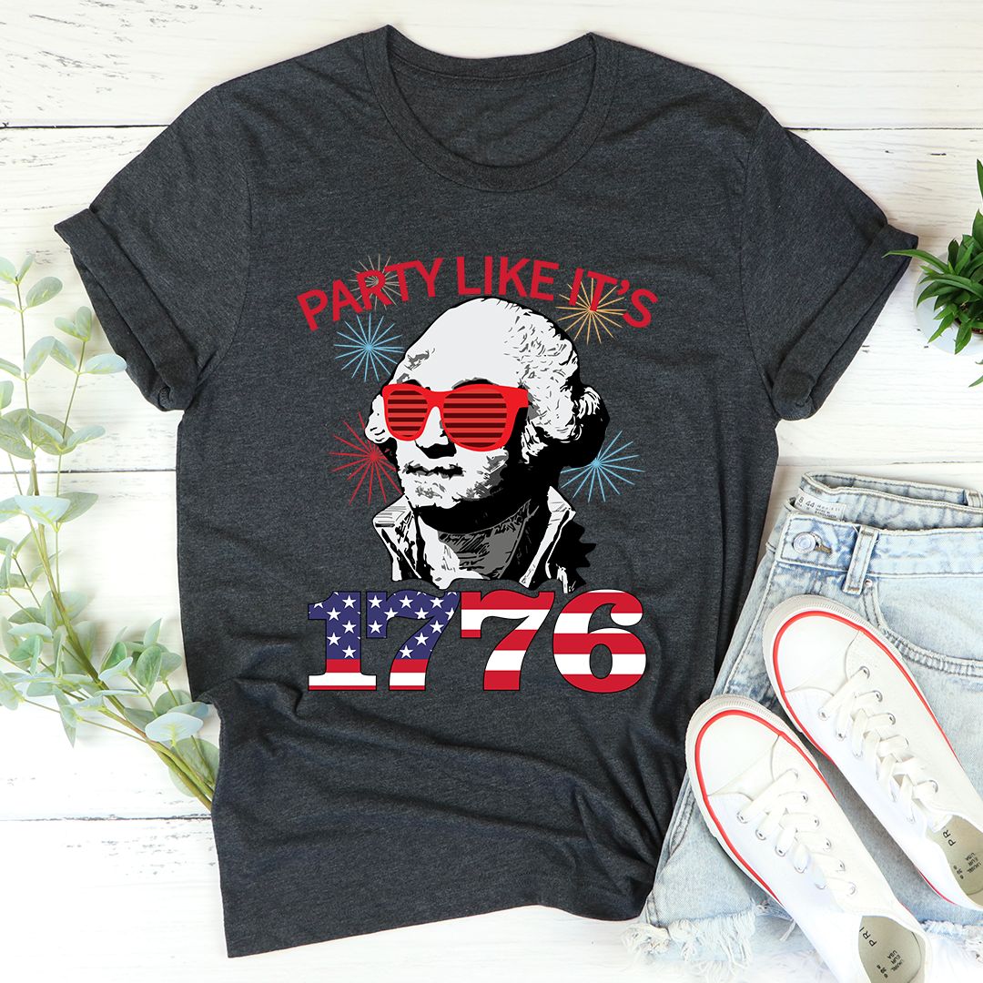 Dark gray t-shirt with a graphic of a historical figure wearing sunglasses and text 'Party Like It's 1776' on a white surface with jeans and shoes.
