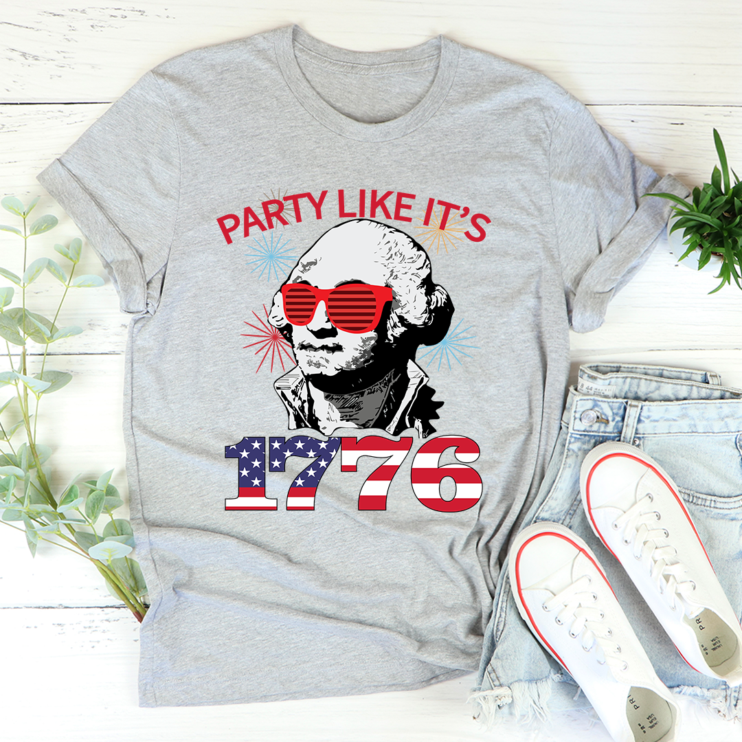 Gray t-shirt with a graphic design featuring a person wearing sunglasses and text 'Party Like It's 1776' on a wooden surface.