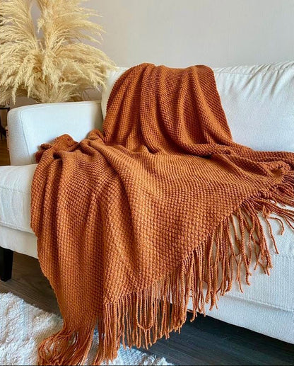 A rust-orange textured knit throw blanket with fringe, draped over a white sofa in a living room setting.