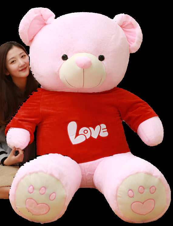Person sitting next to a large pink teddy bear with 'LOVE' on a red shirt in a room with bookshelves.