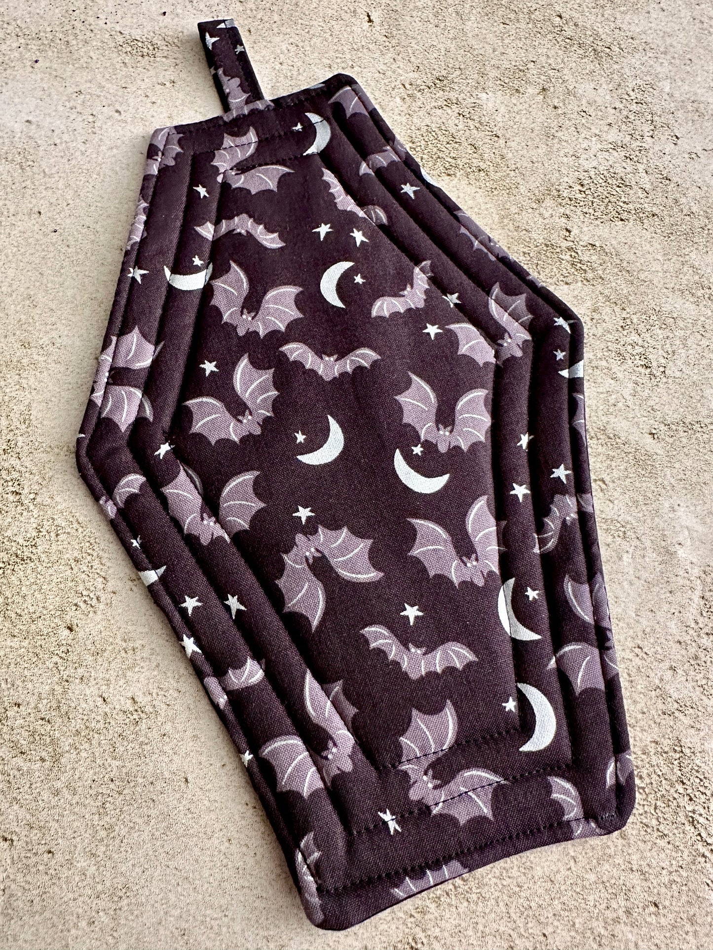Set of two dark purple pot holders with bat and moon pattern on a concrete surface