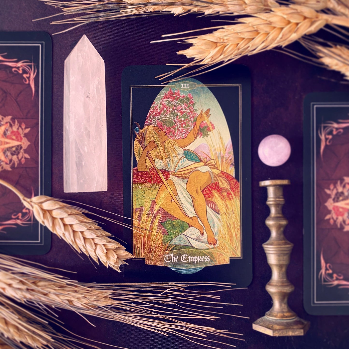 A tarot card set styled on a dark background with decorative wheat stalks, a white crystal point, a pink orb, and a candlestick. The central card is labeled 'The Empress' and depicts an artistic figure seated in a field.