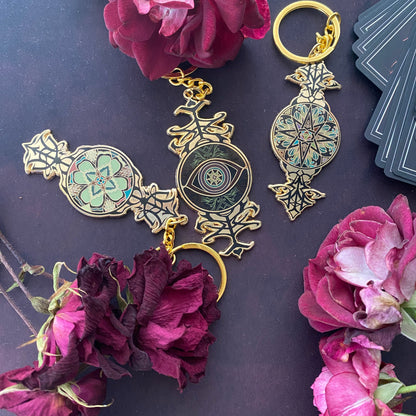 Three ornate gold-toned keychains with intricate floral and eye motifs, styled with dark pink roses on a dark purple background.