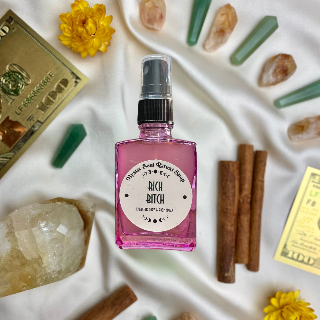 Pink bottle labeled 'Rich Bitch' surrounded by crystals, flowers, and cinnamon sticks on a white fabric background.