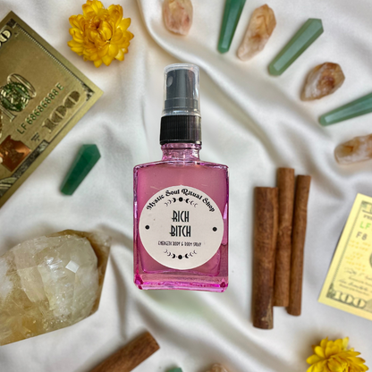 Pink bottle labeled 'Rich Bitch' surrounded by crystals, flowers, and cinnamon sticks on a white fabric background.