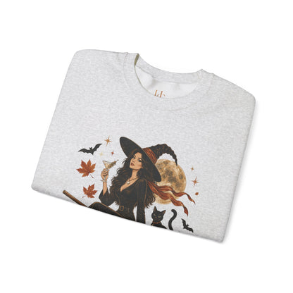 A light heather gray crewneck sweatshirt featuring a graphic print of a witch in a black hat and dress, holding a glass, surrounded by black cats, bats, autumn leaves, and a full moon.