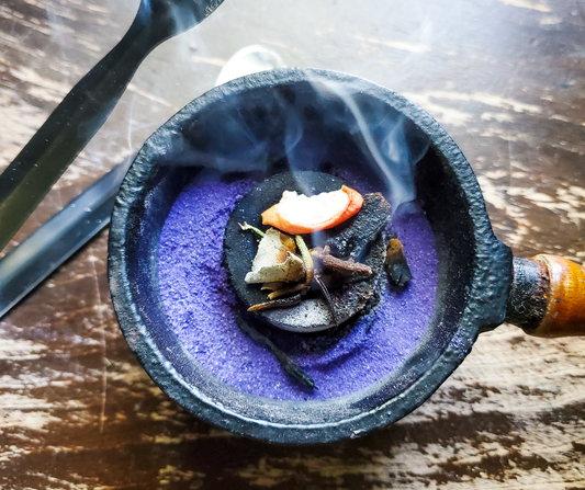Small black pot with purple sand containing incense or similar material on a wooden surface.