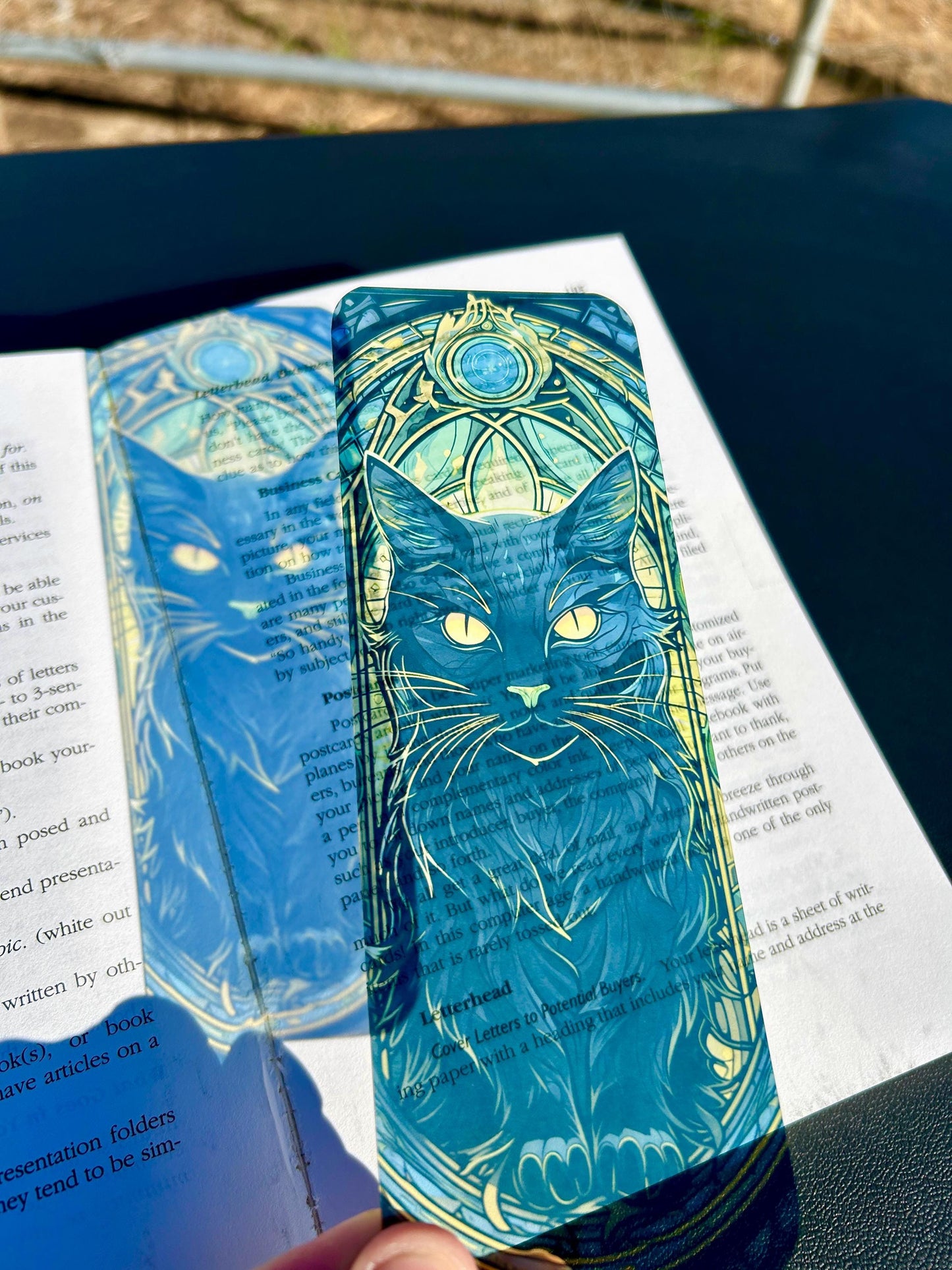 A blue and teal illustrated book bookmark featuring a cat face and ornate decorative border, held in front of an open book on a dark surface outdoors.