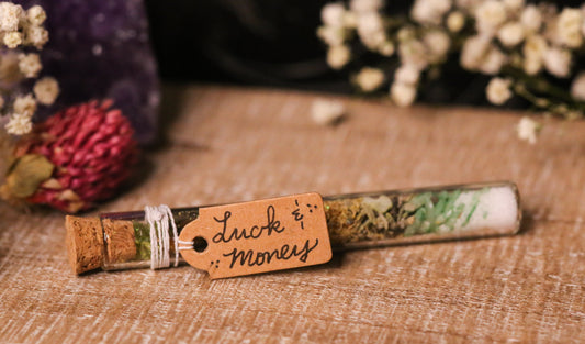 Small glass jar with cork stopper containing herbs, labeled 'Luck & Money', on a textured surface with flowers in the background.