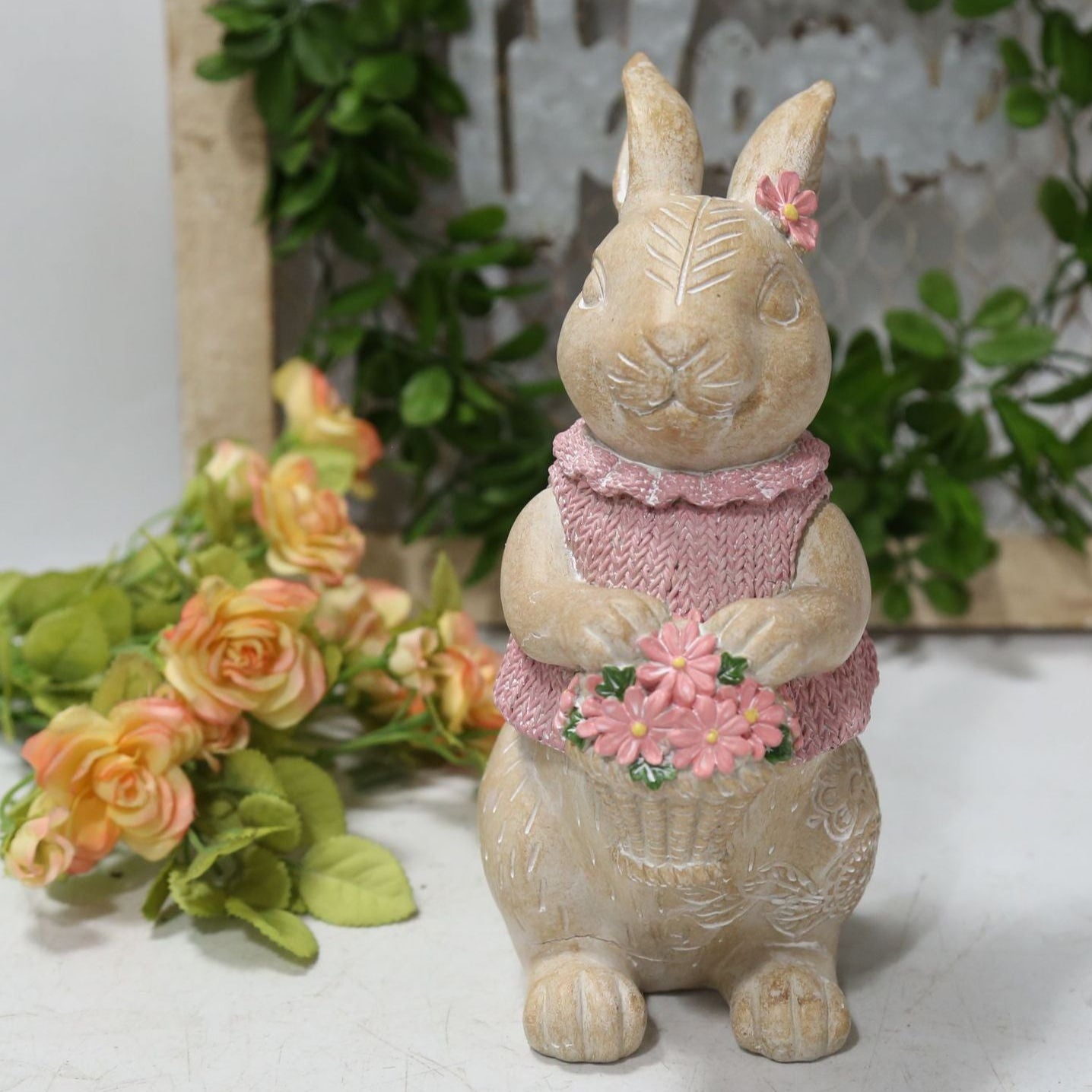 Decorative rabbit figurine with pink outfit and flowers, placed on a white surface with greenery in the background.