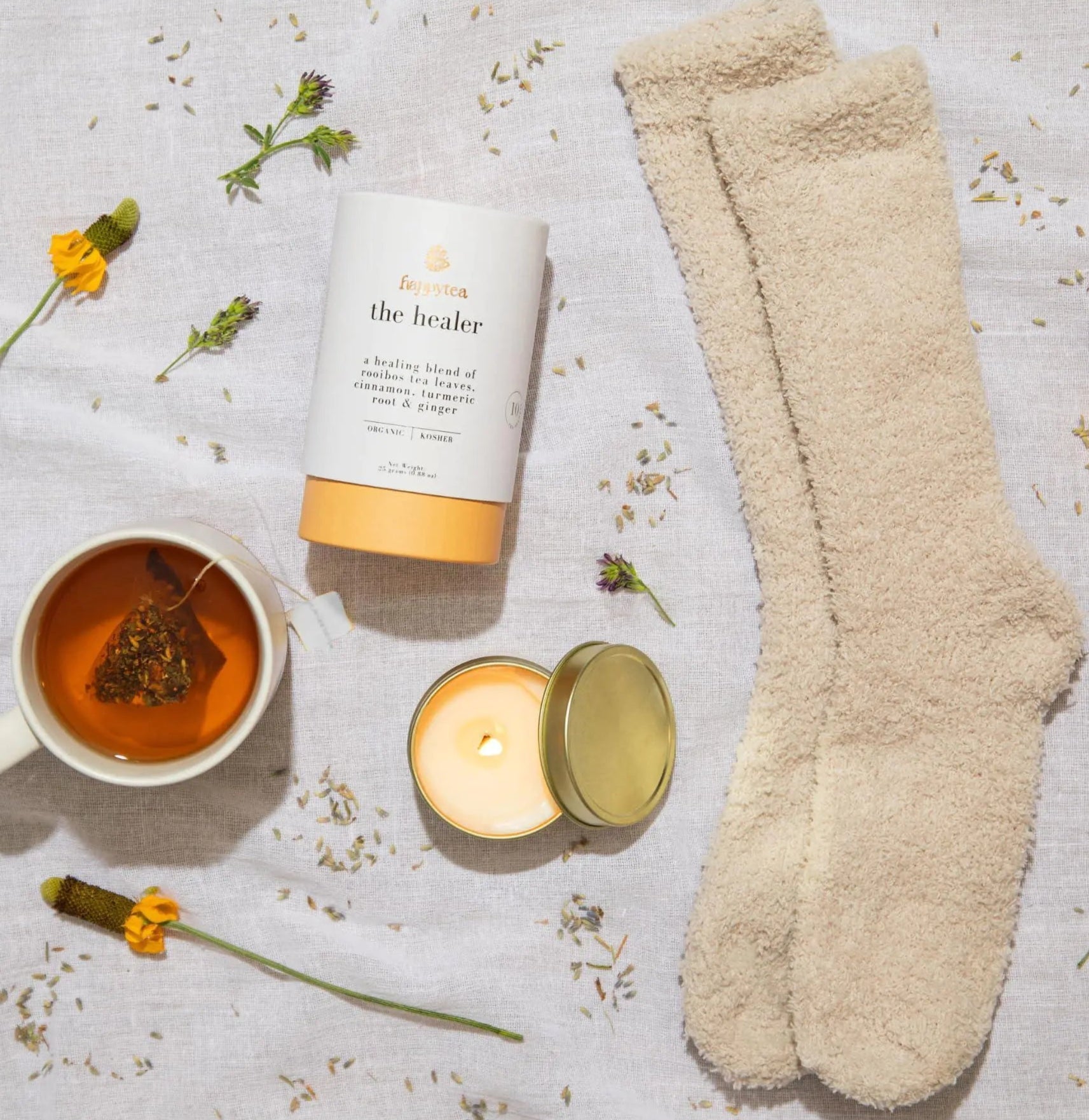 Beige socks, candle, tea cup, and bottle labeled 'The Healer' on a textured surface with flowers.