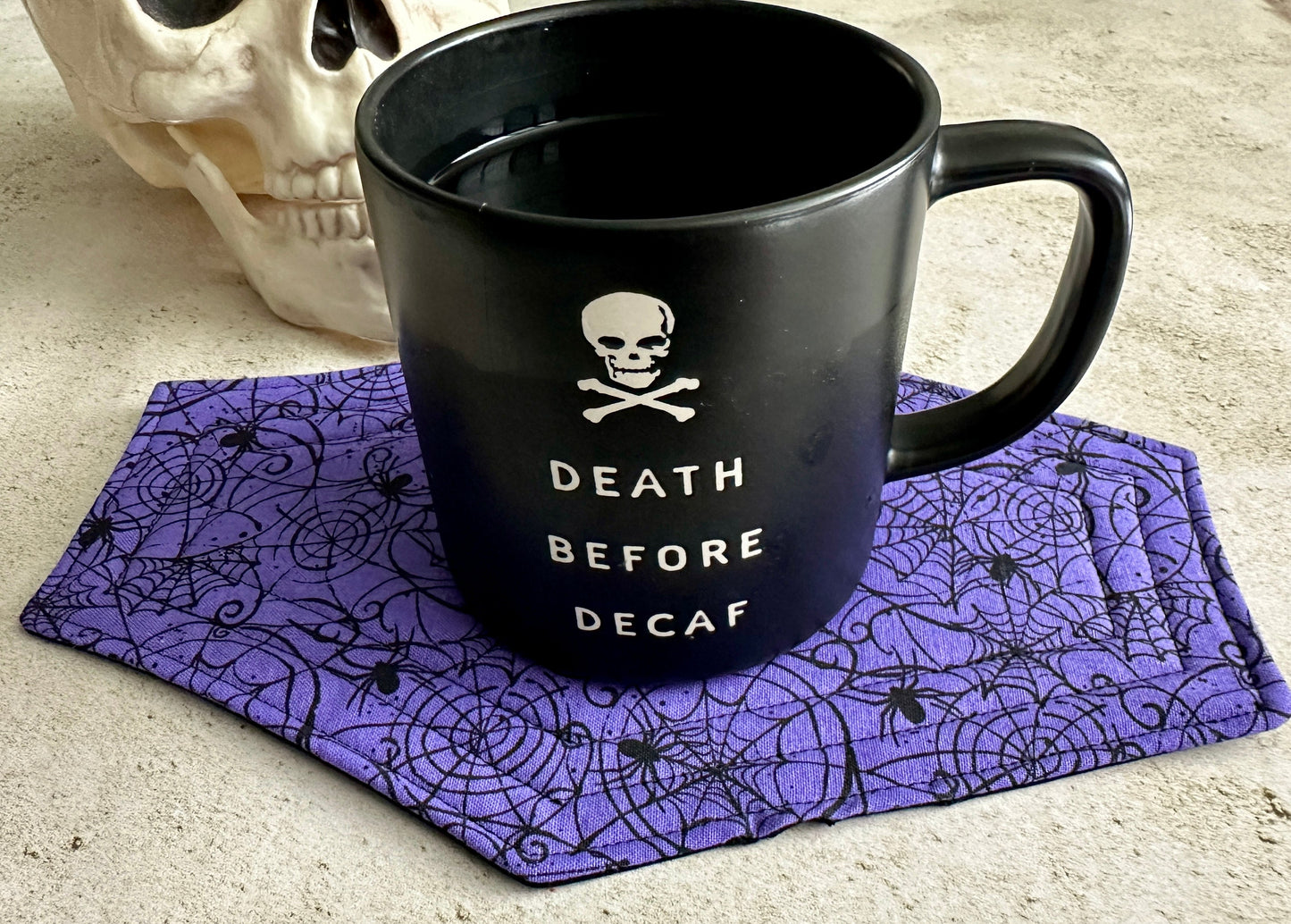Black mug with 'Death Before Decaf' text on a purple spiderweb coaster, next to a skull.