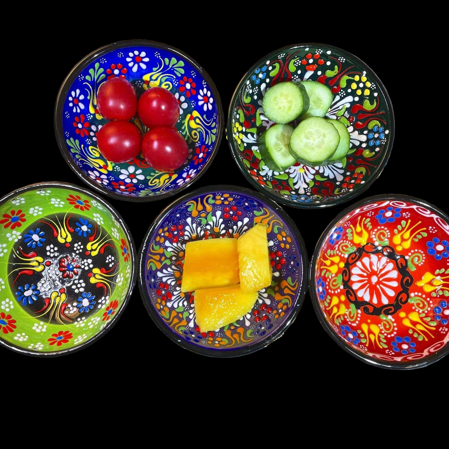 Five colorful plates with floral patterns containing red tomatoes, green cucumbers, and yellow pineapple chunks on a decorative surface.