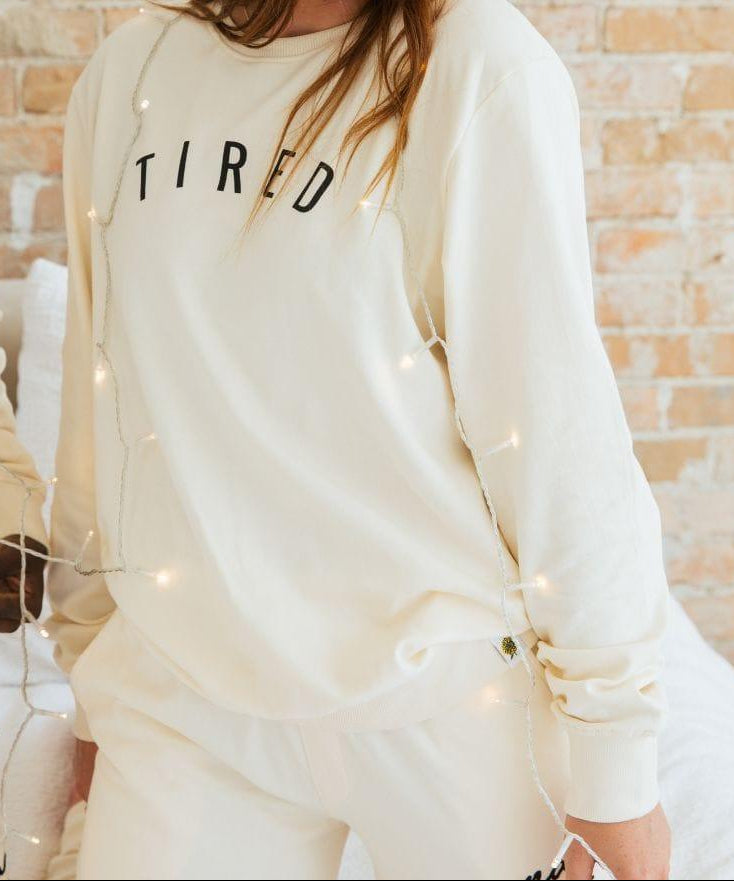Woman wearing a cream-colored sweatshirt with 'TIRED' text, standing against a brick wall.