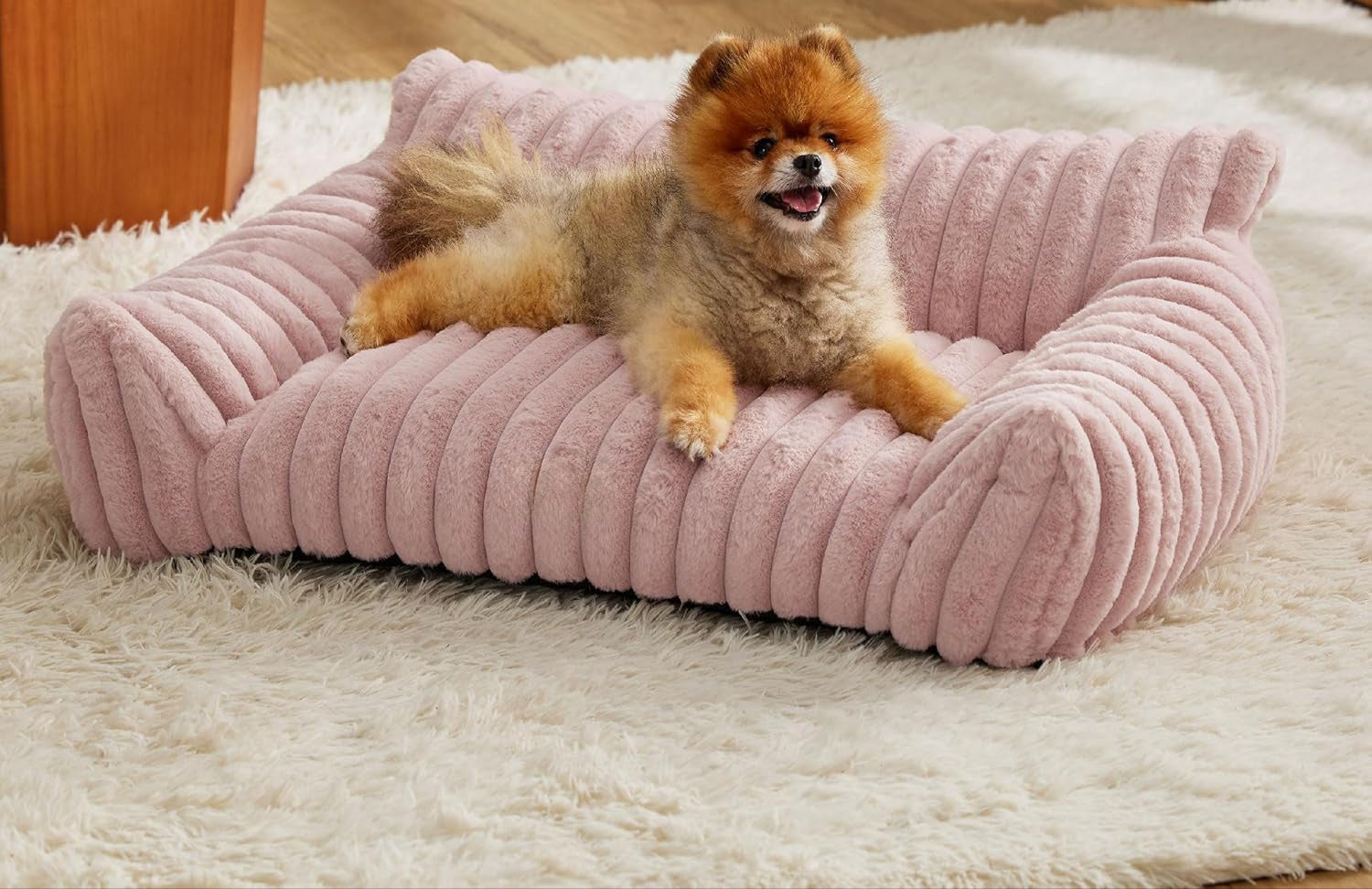 Small dog on a pink plush dog bed in a cozy room with a fireplace.