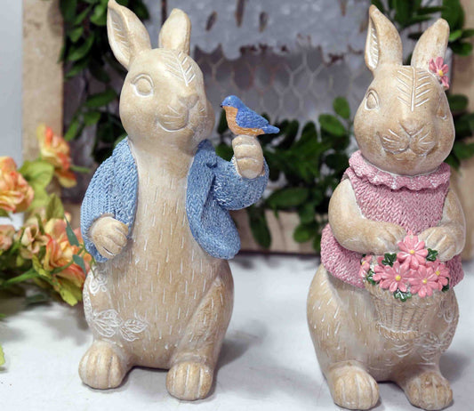 Two decorative rabbit figurines, one with a blue scarf and bird, the other with a pink dress and flowers, against a neutral background.