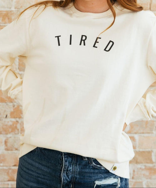 Person wearing a cream sweatshirt with 'TIRED' text against a brick wall.