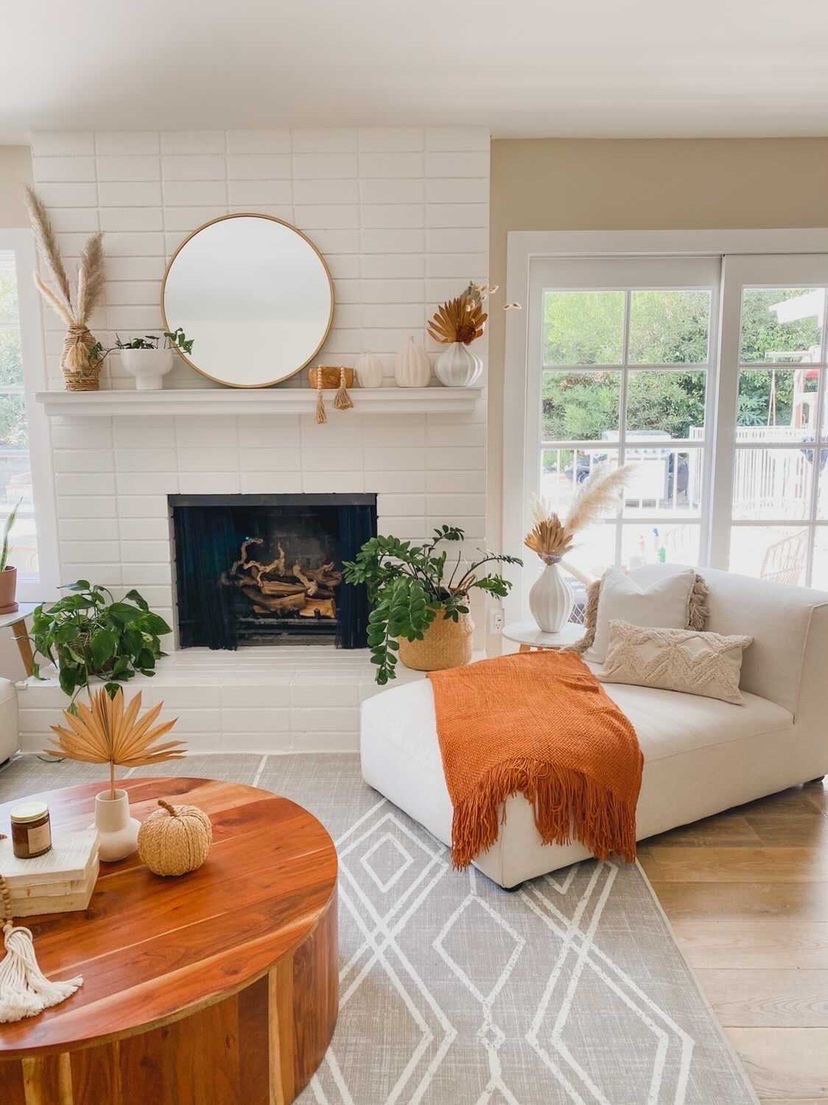 A bright, modern living room featuring a white brick fireplace with a white mantel decorated with autumn-themed items, a white chaise lounge with an orange throw blanket, a round wooden coffee table, and a gray geometric rug.
