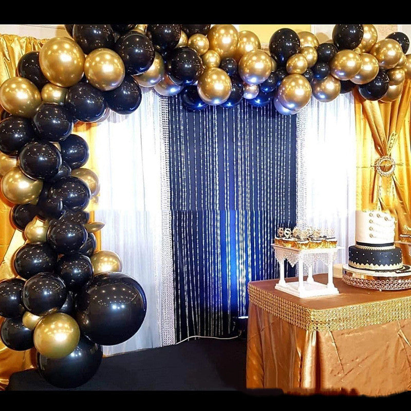 Decorative arch of black and gold balloons with a cake on a table in the background.