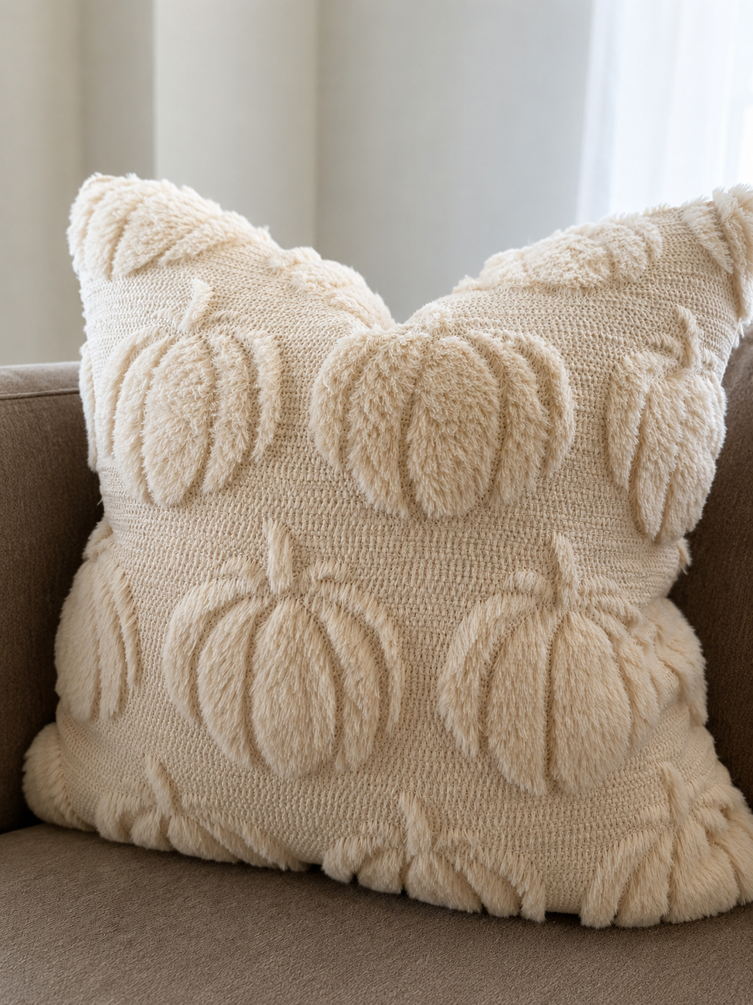A square cream throw pillow with a textured, fuzzy knit design featuring raised pumpkins and leaves, resting on a brown sofa.