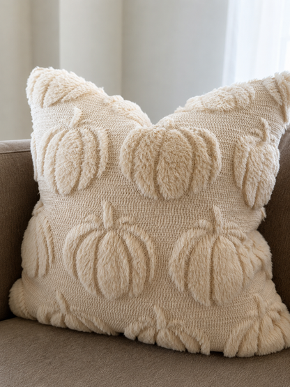 A square cream throw pillow with a textured, fuzzy knit design featuring raised pumpkins and leaves, resting on a brown sofa.