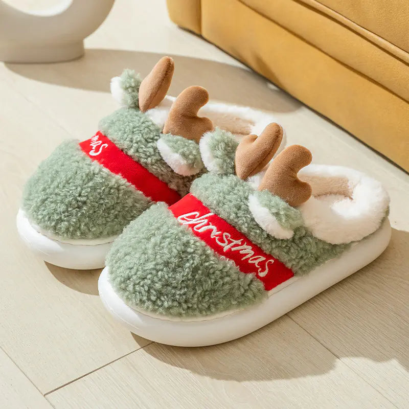 Green Christmas-themed slippers with reindeer ears and a red band on a light wooden floor.