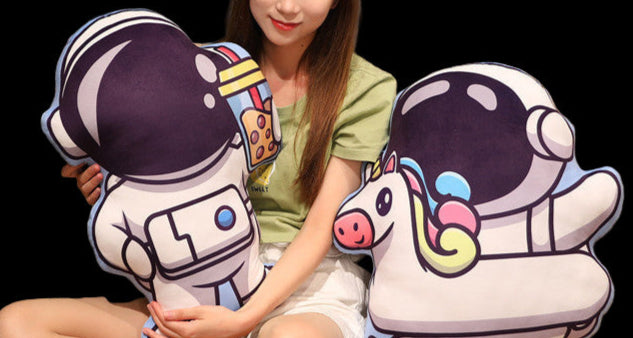 Woman sitting on a couch holding two plush toys shaped like astronauts with unicorn helmets.