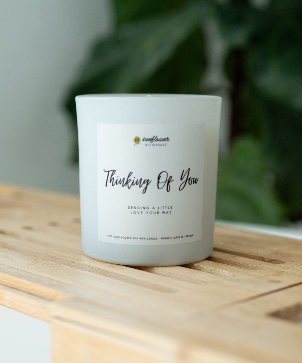 Candle with 'Thinking Of You' text on a wooden surface with a blurred green plant background