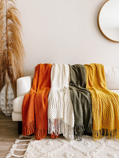 Four textured knit throw blankets in orange, cream, olive green, and mustard yellow draped over a white sofa in a styled living room setting.
