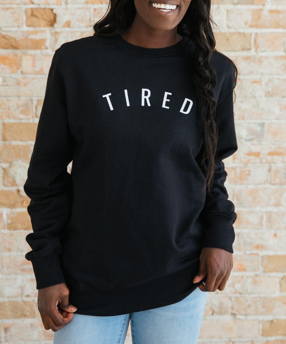 Person wearing a black sweatshirt with 'TIRED' text against a brick wall.