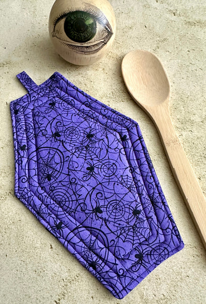 Purple oven mitt with black spider design on a beige surface with a wooden spoon and eye-shaped object.