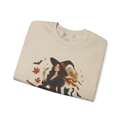 A folded cream-colored crewneck sweatshirt featuring a detailed graphic of a witch in a black hat and dress, holding a martini glass, surrounded by autumn leaves, bats, a black cat, and a full moon.