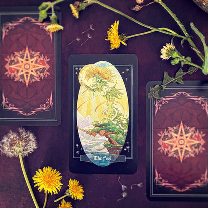 A tarot card deck styled flat-lay on a dark purple surface, featuring 'The Fool' card in the center with yellow flowers and dandelions scattered around it.
