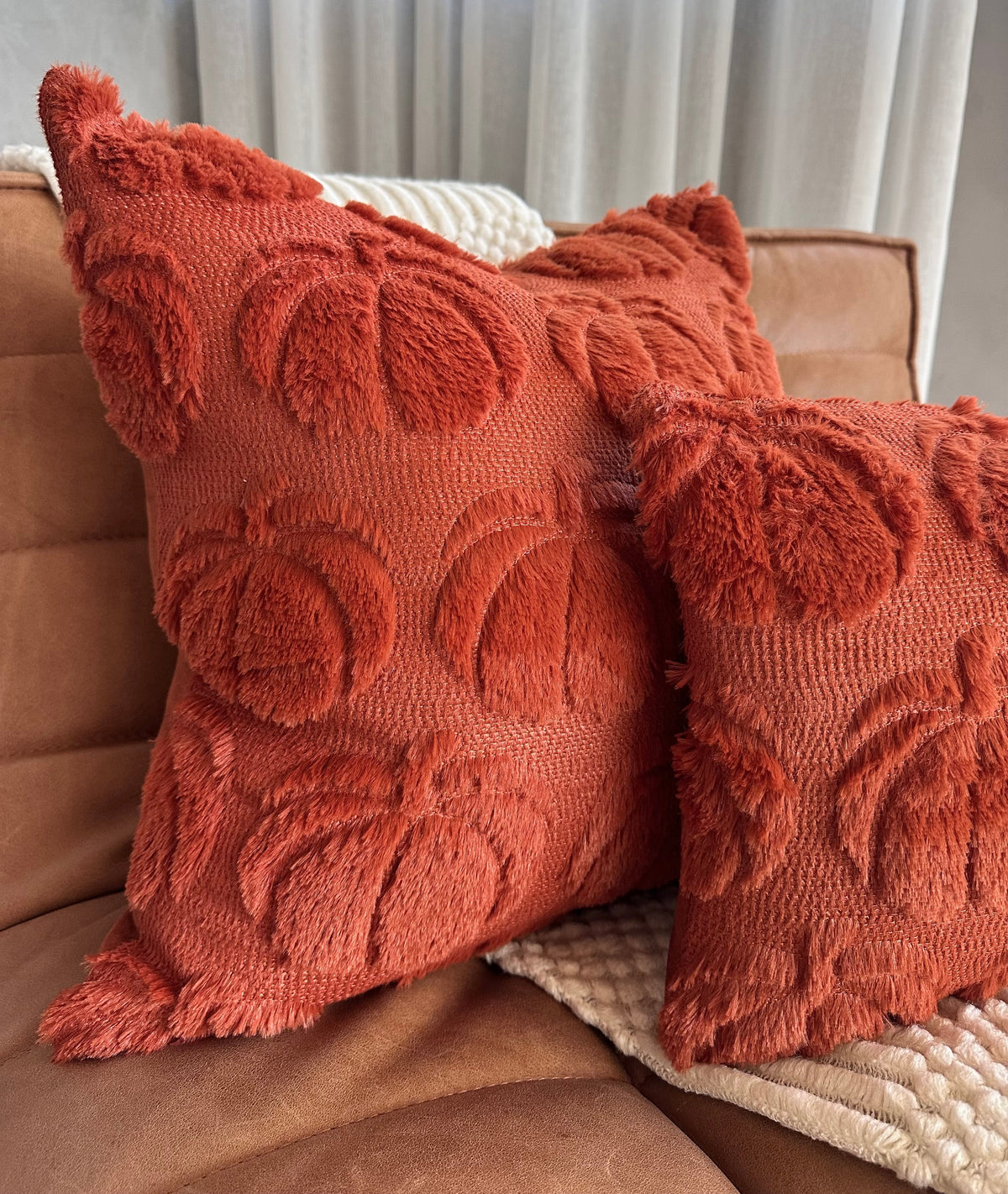 Two textured orange pillows on a brown couch with light curtains in the background.