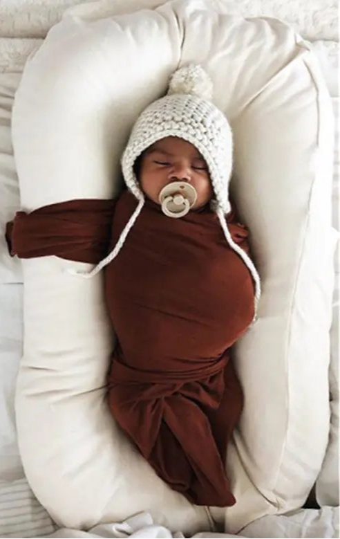Baby wrapped in brown swaddle with white knit hat and pacifier, lying on a white cushion.