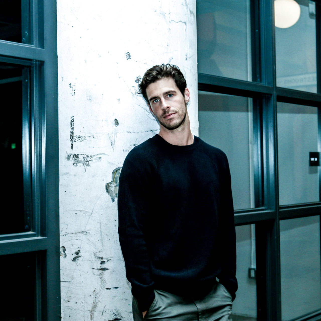 Man in a black sweater leaning against a textured wall with large windows.