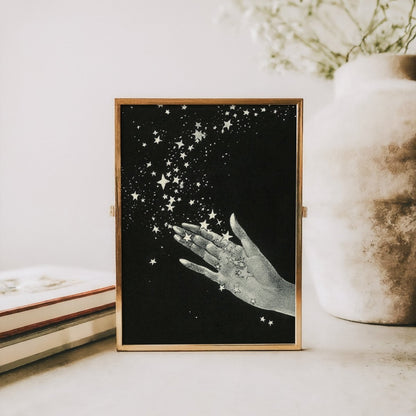 Framed artwork of a hand holding stars on a table with books and a vase.