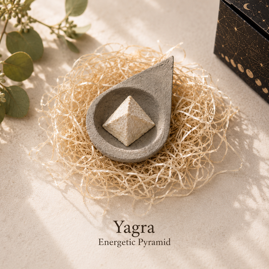 A teardrop-shaped gray stone holder containing a small beige yagara crystal pyramid, styled on a bed of natural jute fiber with eucalyptus leaves and a dark decorative box in the background. Text at the bottom reads 'Yagra Energetic Pyramid'.