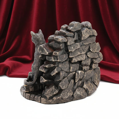 A decorative figurine of a black cat sitting on a rustic stone wall, crafted with a textured metallic bronze finish, set against a plain white background.