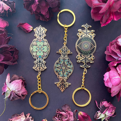 Three ornate metal keychains with gold-tone chains and rings, decorated with intricate floral and eye motifs in gold, black, and turquoise, arranged on a dark background with dried rose petals.